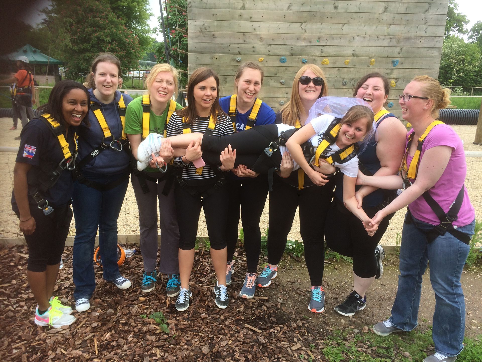 Group of women in harnesses pose for photo near climbing wall, some holding up another.