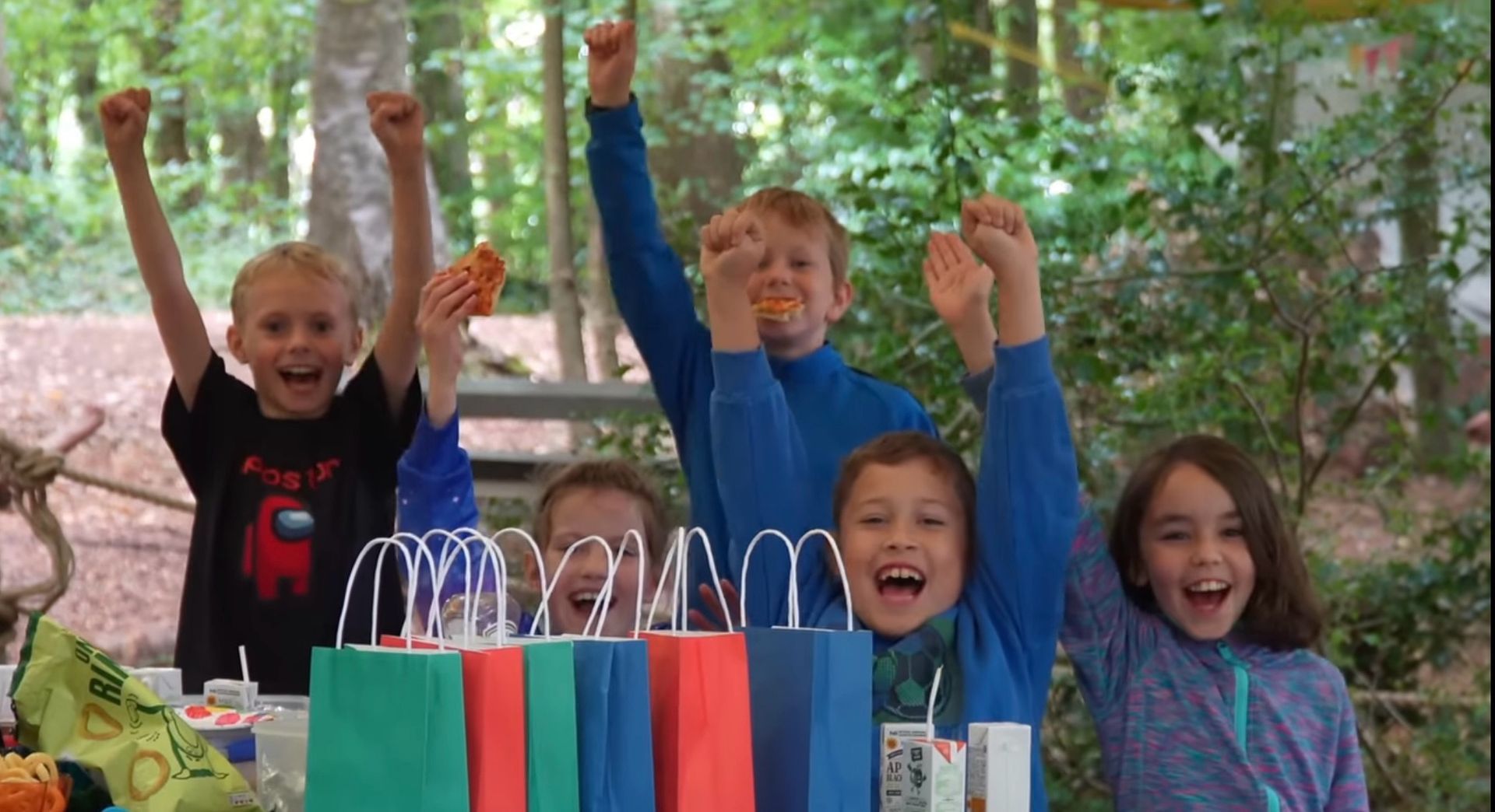 Children cheering with arms raised, celebrating behind colorful gift bags; outdoor setting.Having a Birthday Party Tea