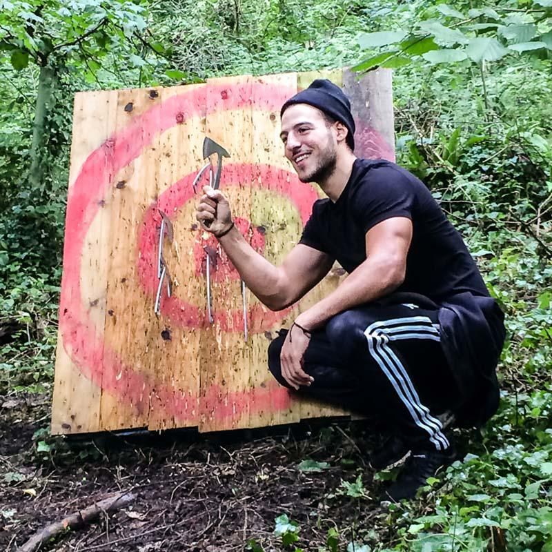 Man squats, holding axe in front of wooden target with axe imbedded. Forest setting.