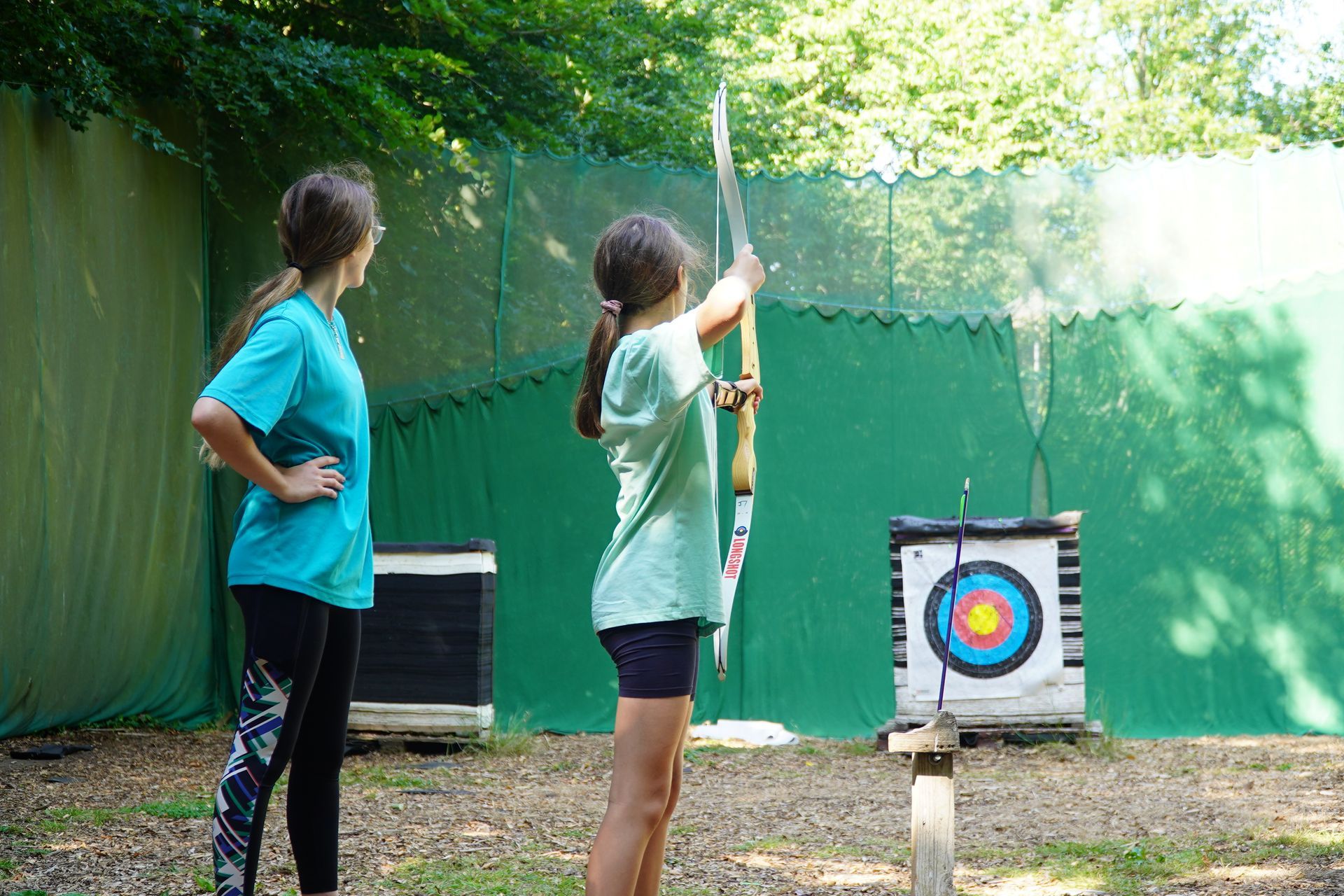Girl aims bow and arrow at target, another watches. Green backdrop, sunny outdoor setting.