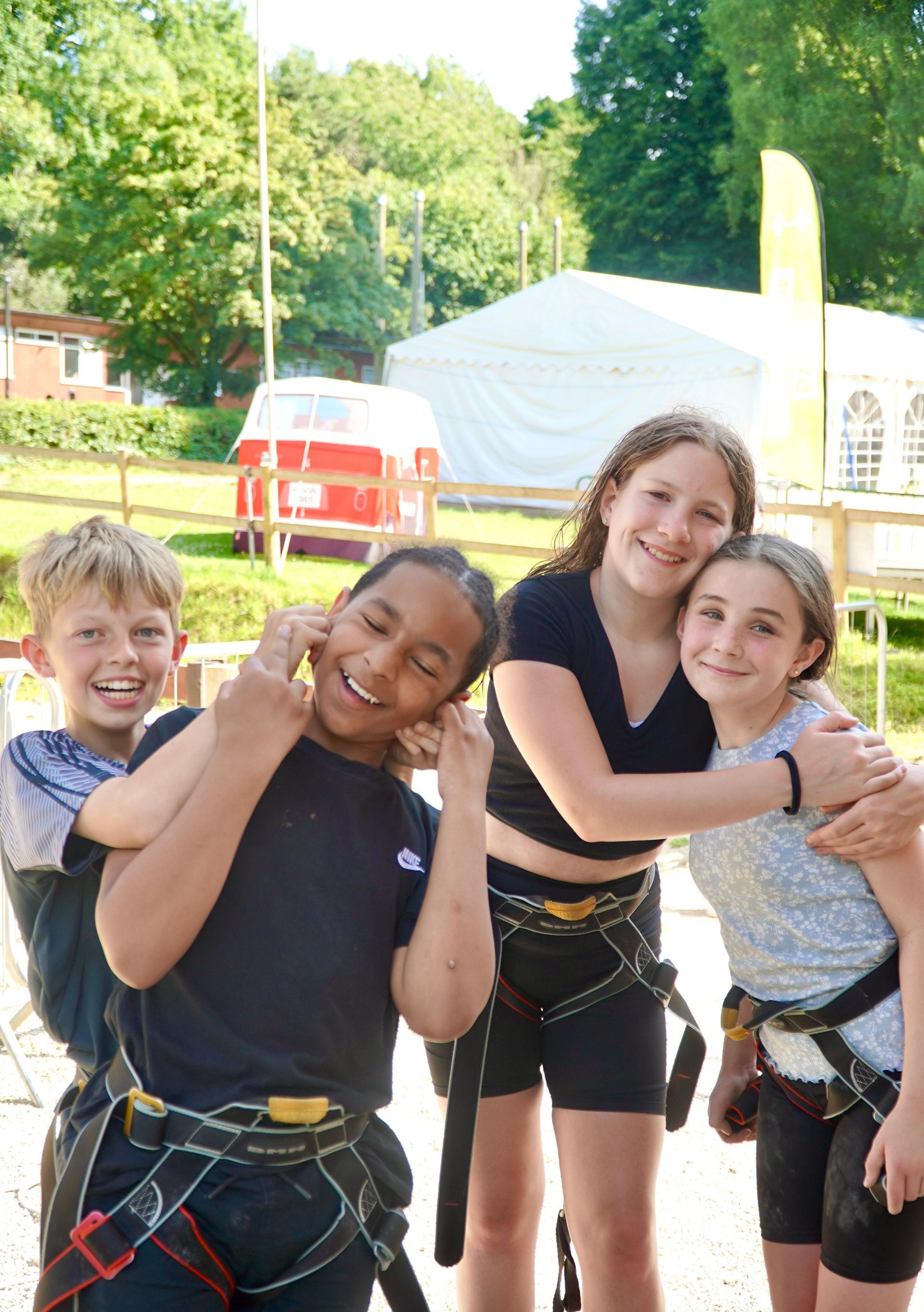 Four kids in harnesses smile near a white tent and red van; they embrace.