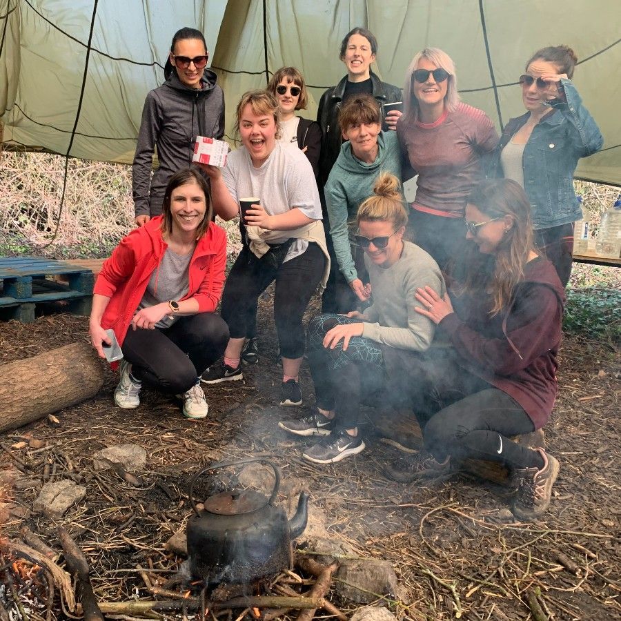 Group of women smiling around a campfire; kettle on fire, outdoors under a tarp.