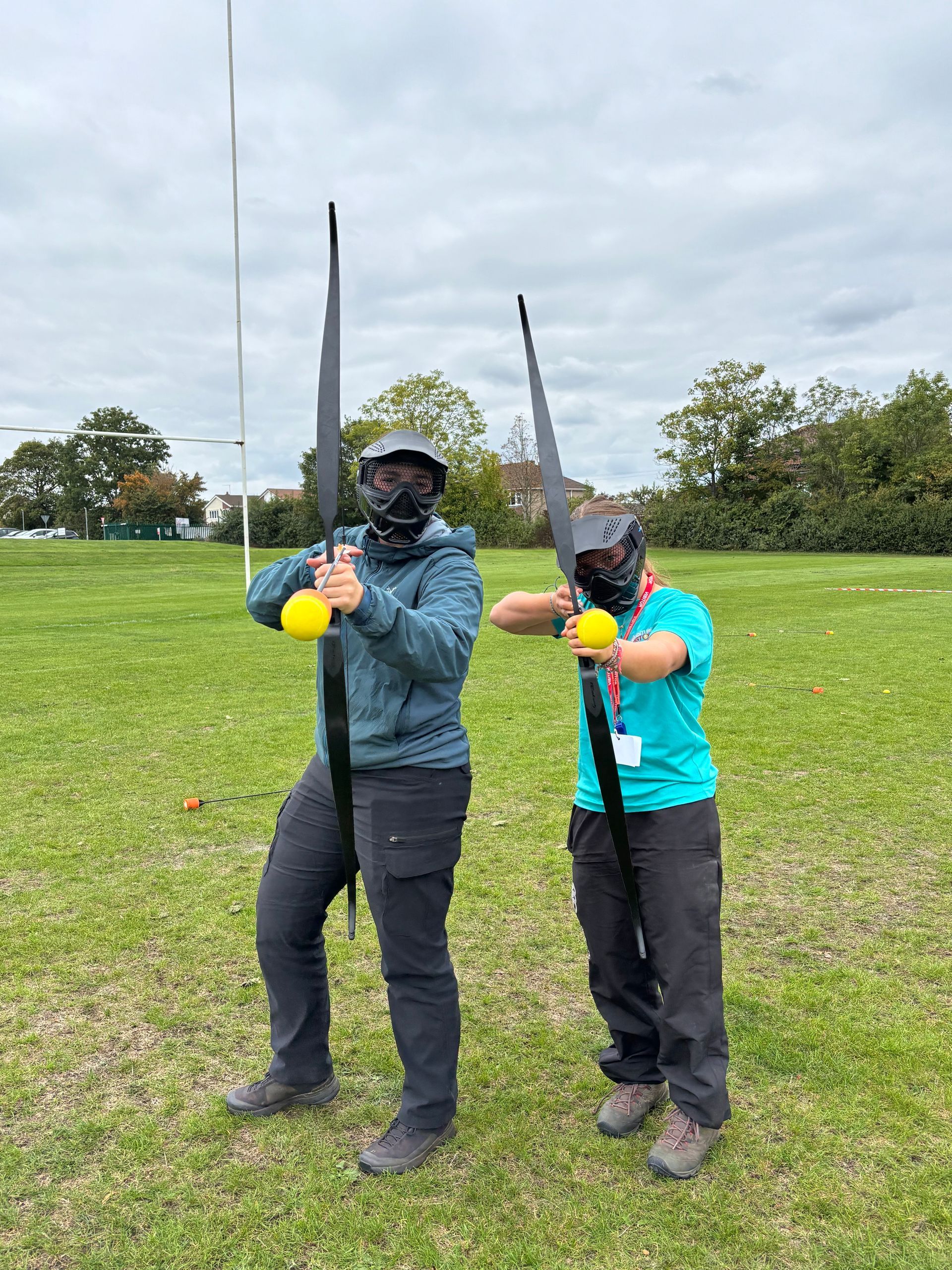 Two people in helmets and masks hold large black arrows with yellow tips outdoors.