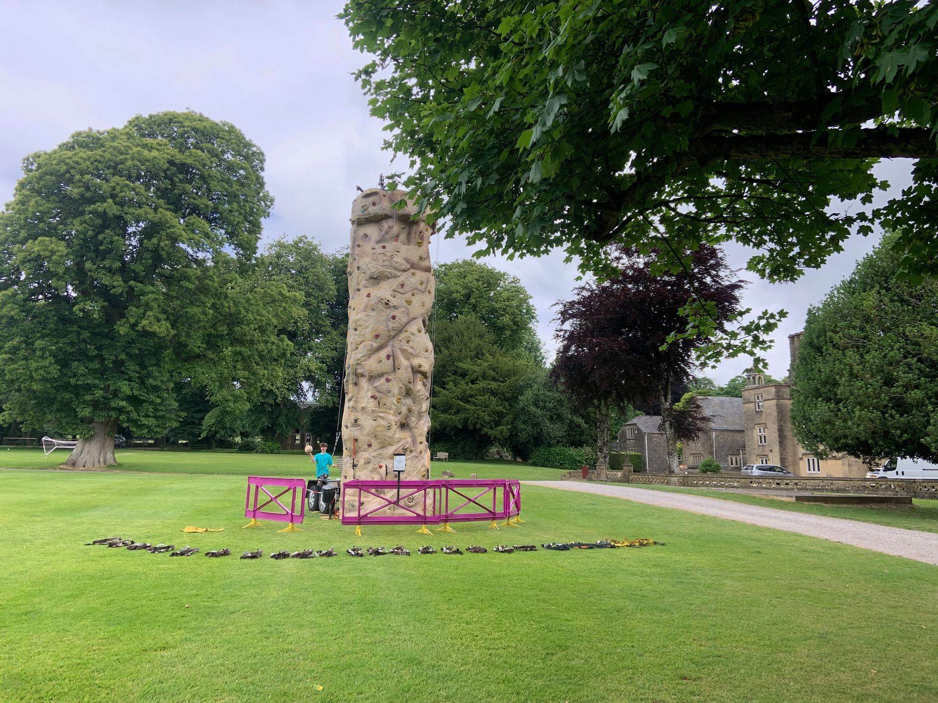 Rock climbing wall on a grassy lawn. Person in green shirt prepares to climb; trees and building in the background.