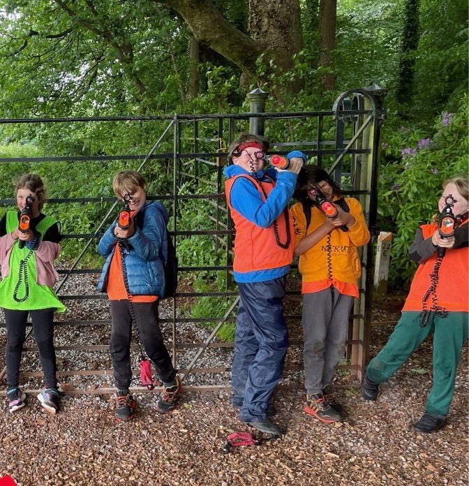 Children aiming toy guns, wearing orange vests. Standing in front of a fence, green foliage in the background.
