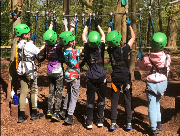 Children in green helmets, harnessed, reaching for ropes on a treetop adventure course.
