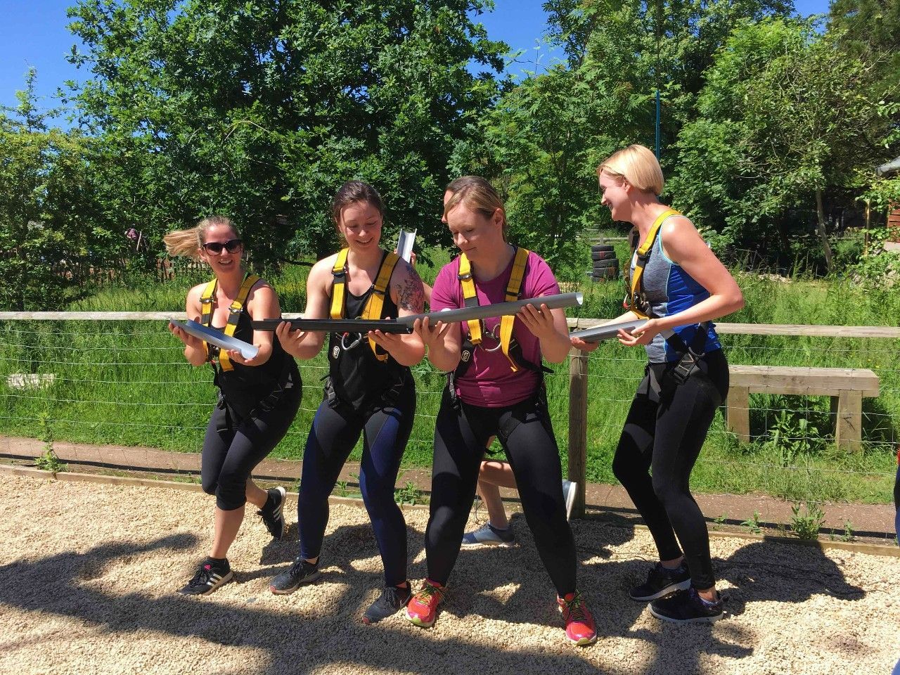 Four women in harnesses holding metal bars, outdoors near a fence, trees in background.