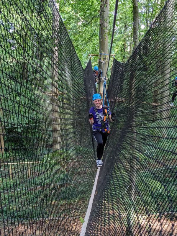 People on a high ropes course, walking across a narrow beam. Black netting on sides, trees in background.