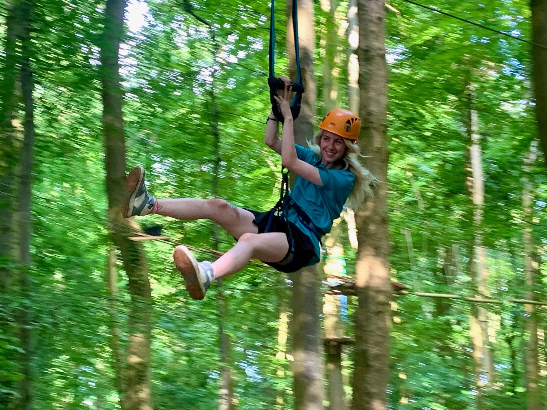 Woman ziplining through trees, wearing a helmet and harness, smiling. zip wire