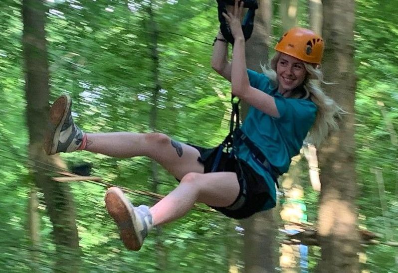 Woman on a zip line in a forest, wearing a helmet and harness, smiling.