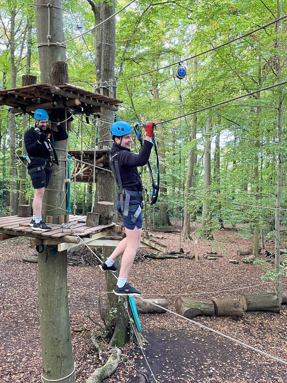 Two people in helmets and harnesses on a treetop adventure course, forest setting. One person zips down a wire.