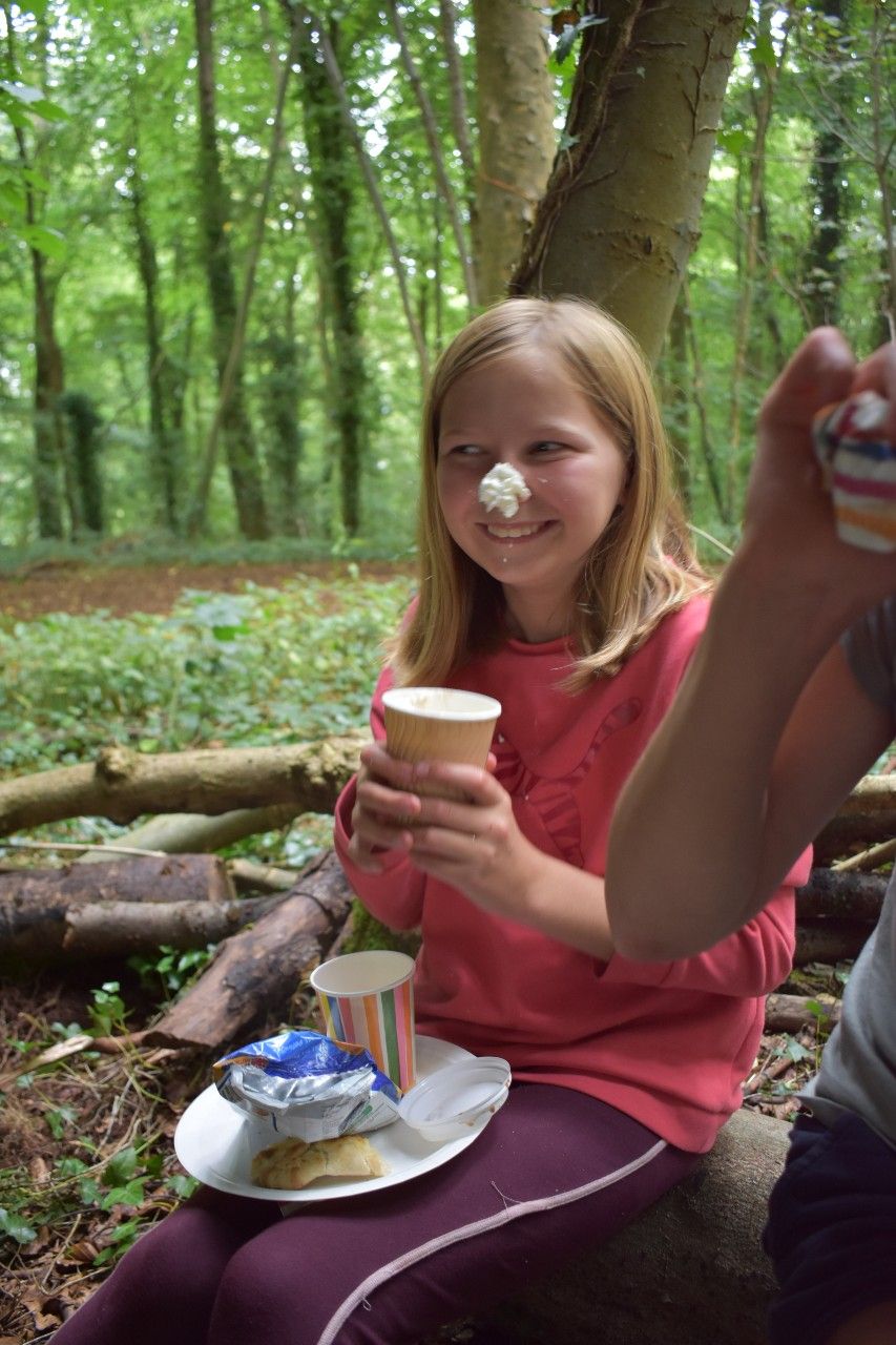 Girl with whipped cream on her nose, sitting in a forest, smiling while holding a cup.