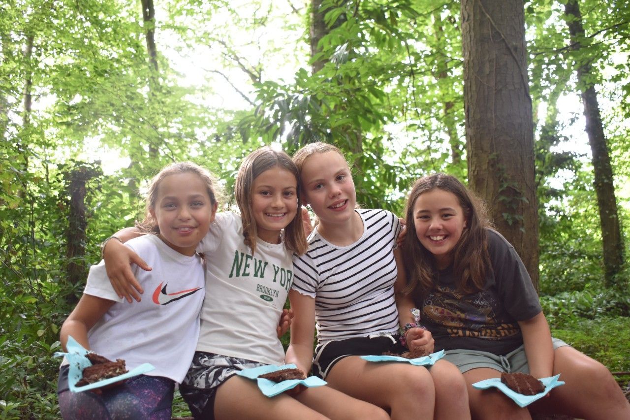 Four smiling girls with treats sit together outdoors in a forest.