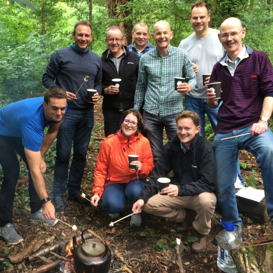 Group of people roasting marshmallows around a campfire, smiling, holding mugs, in a wooded area.