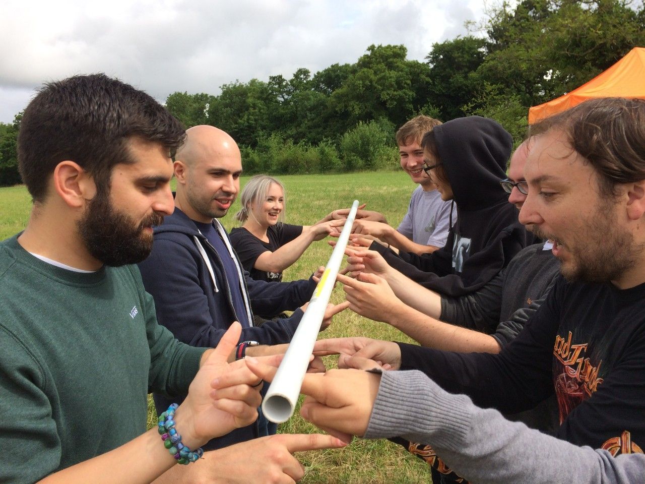 Group of people outdoors, holding a long, white pipe with hands. They point at the pipe with concentrated expressions.