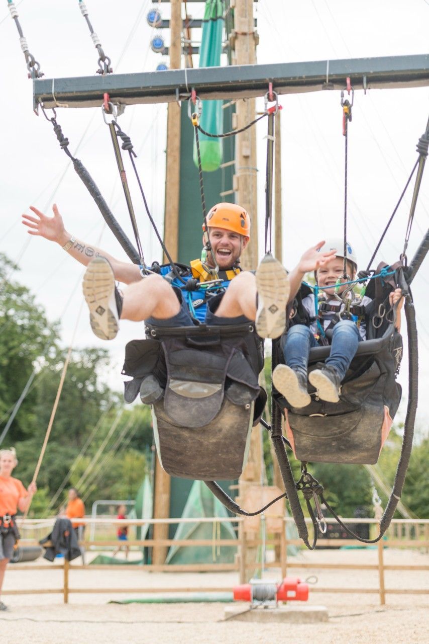 Man and child in harnesses, swinging on an adventure course. They wave and smile. Orange and green background.