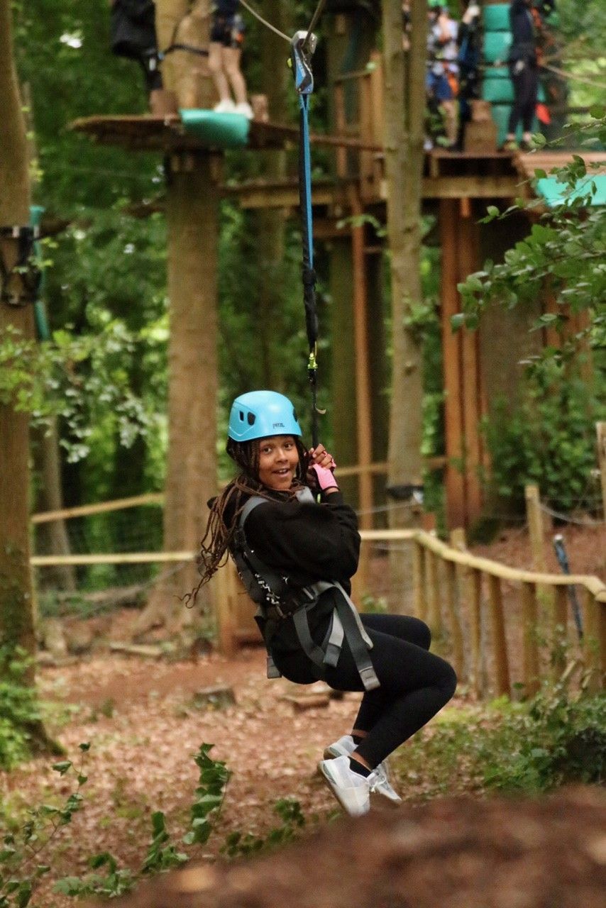 Girl on zip line in forest, wearing blue helmet, smiling, black clothing, adventure park setting. zip wire near me