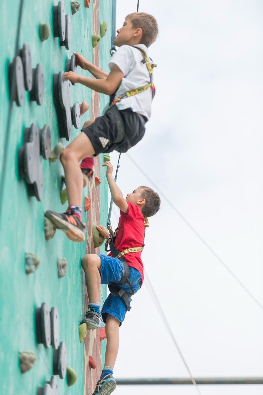 Two boys climbing a green outdoor climbing wall, wearing harnesses, under a cloudy sky.