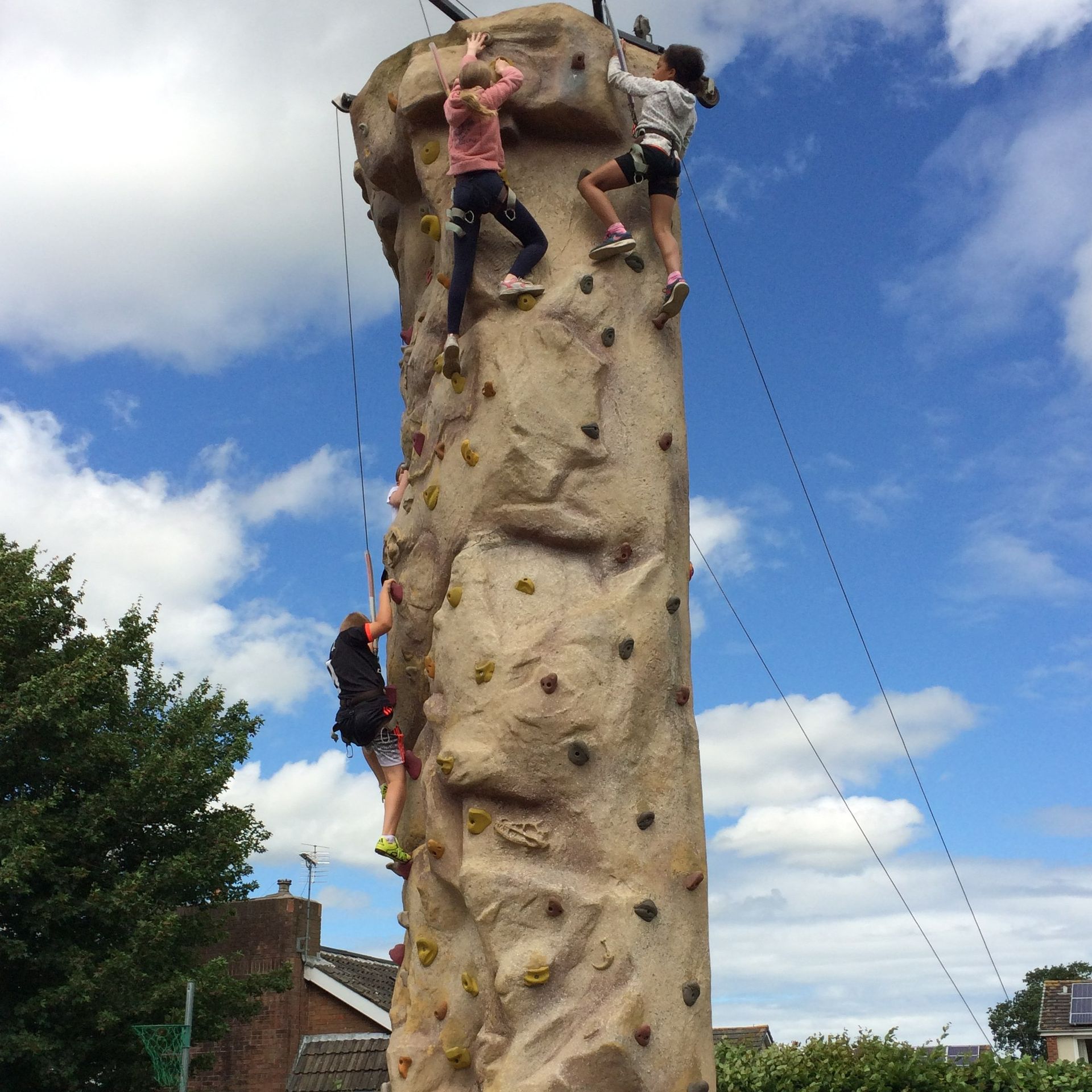 Three people climb a tall rock climbing wall outdoors under a blue sky with clouds.