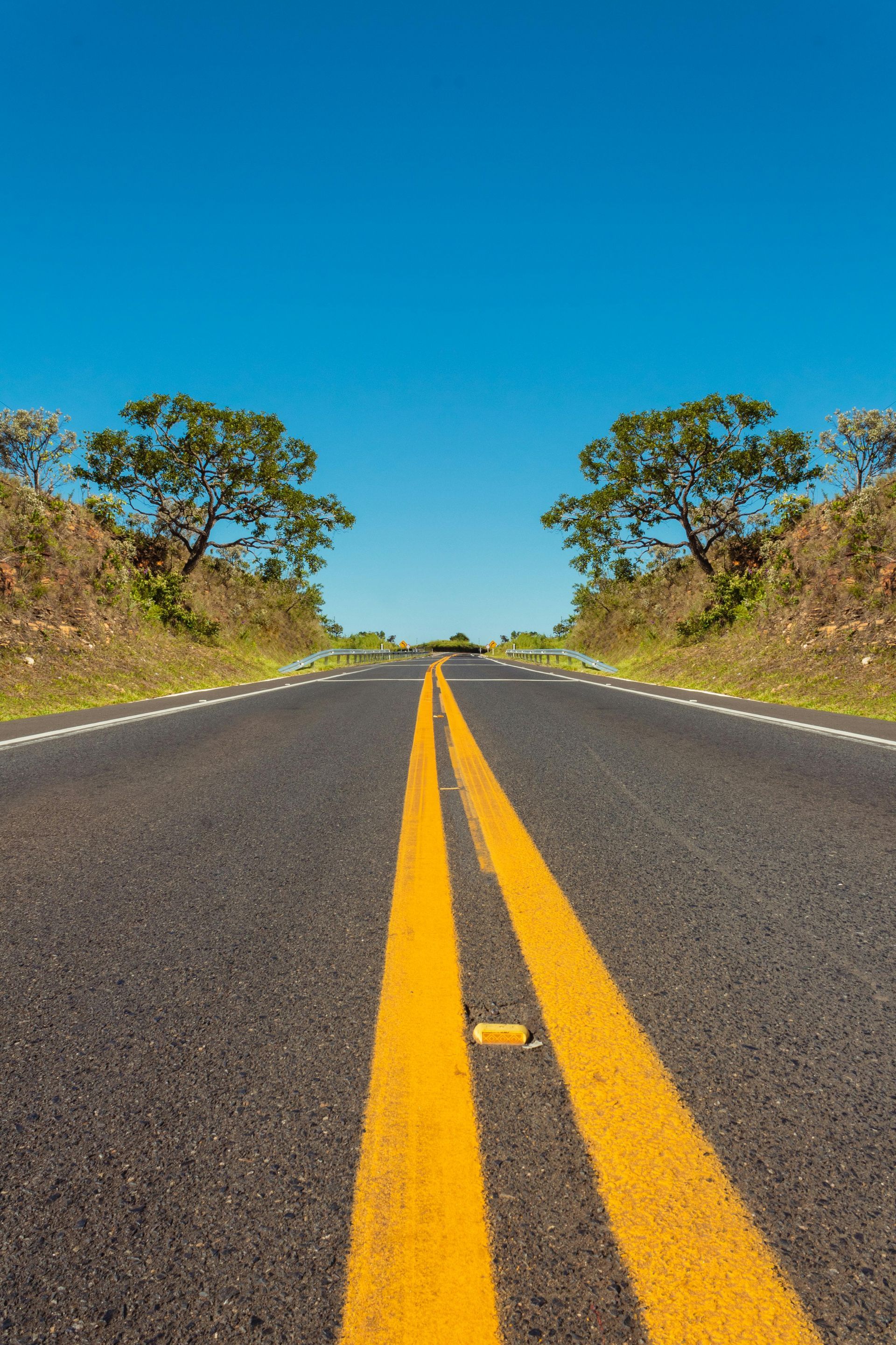 Uma estrada asfaltada com faixas amarelas duplas estende-se em direção a um céu azul claro entre colinas gramadas e arborizadas.