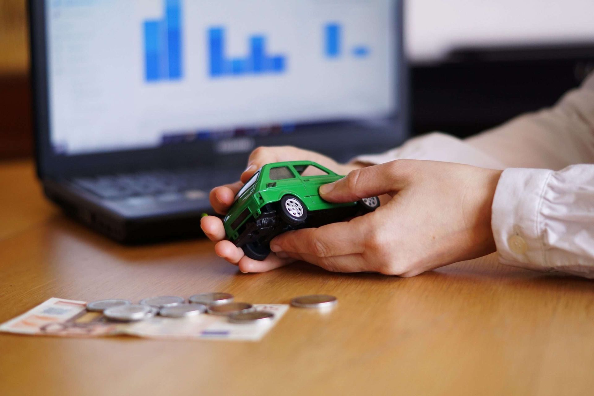 Mãos seguram um pequeno carrinho de brinquedo verde em uma mesa com um laptop exibindo gráficos e algumas moedas espalhadas.