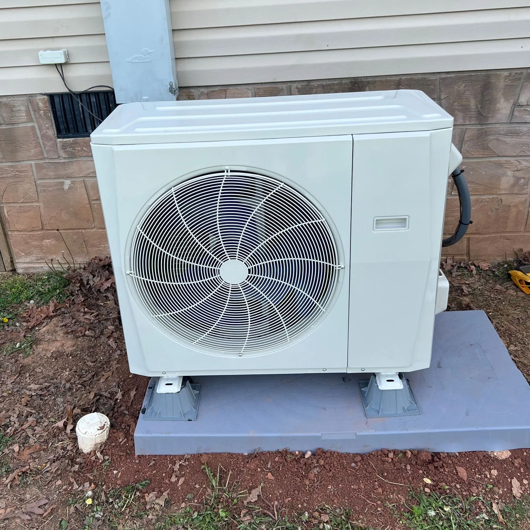 White outdoor air-conditioning unit with a large fan, mounted beside a house on a concrete pad.