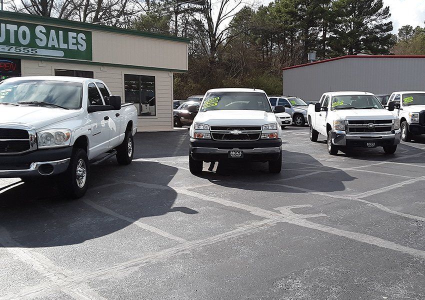 Cars for sale at a car dealership, including white pickup trucks, gray pavement, and a building with 