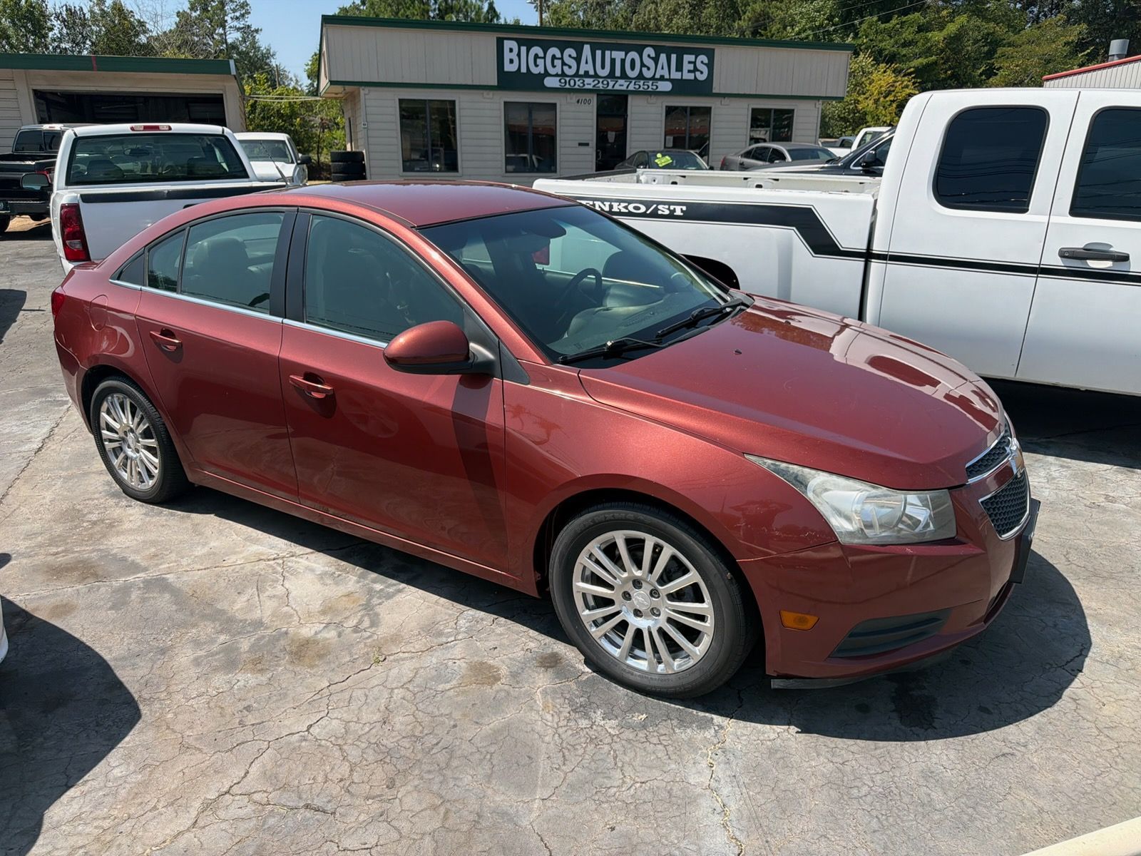 Red sedan parked in a lot in front of a Big3 Auto Sales building, with white trucks nearby