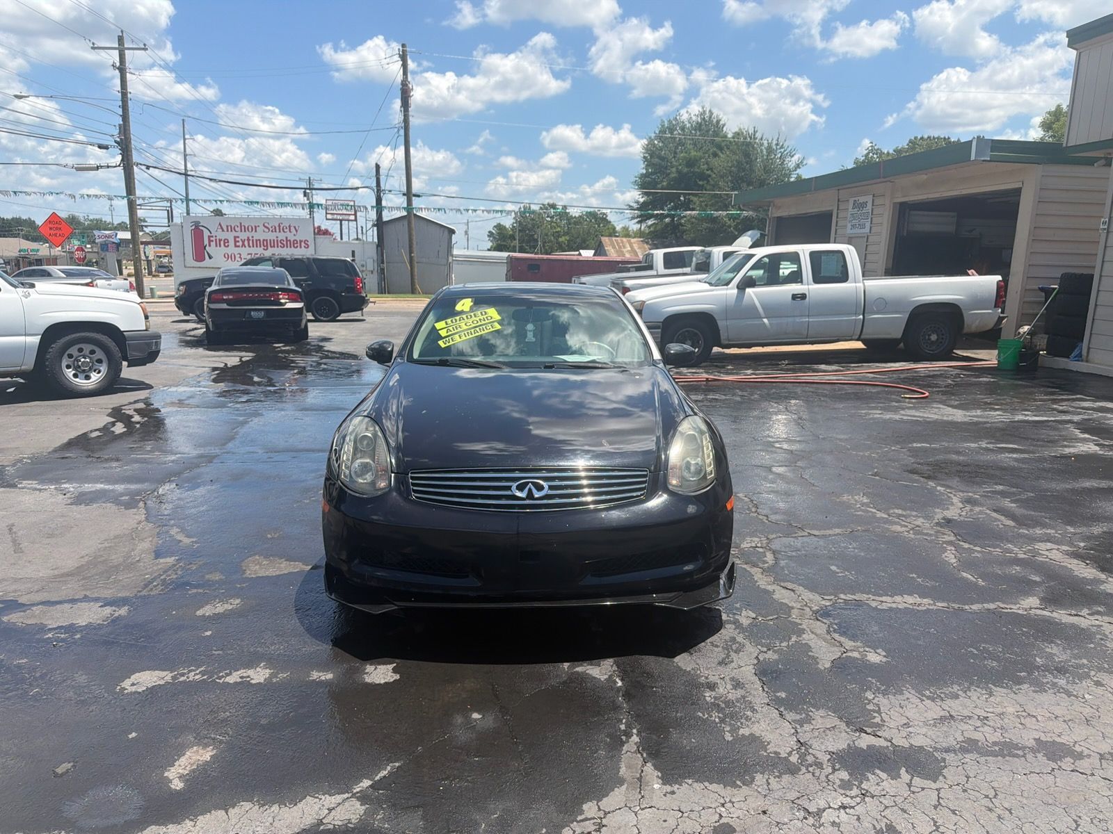 Black car parked on a wet lot with trucks and buildings in the background under a partly cloudy sky