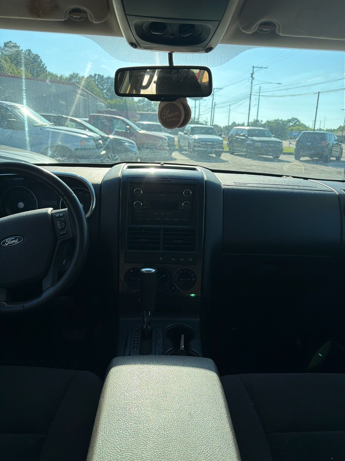 Car interior view with dashboard and steering wheel, parked in a sunny lot with cars visible ahead.