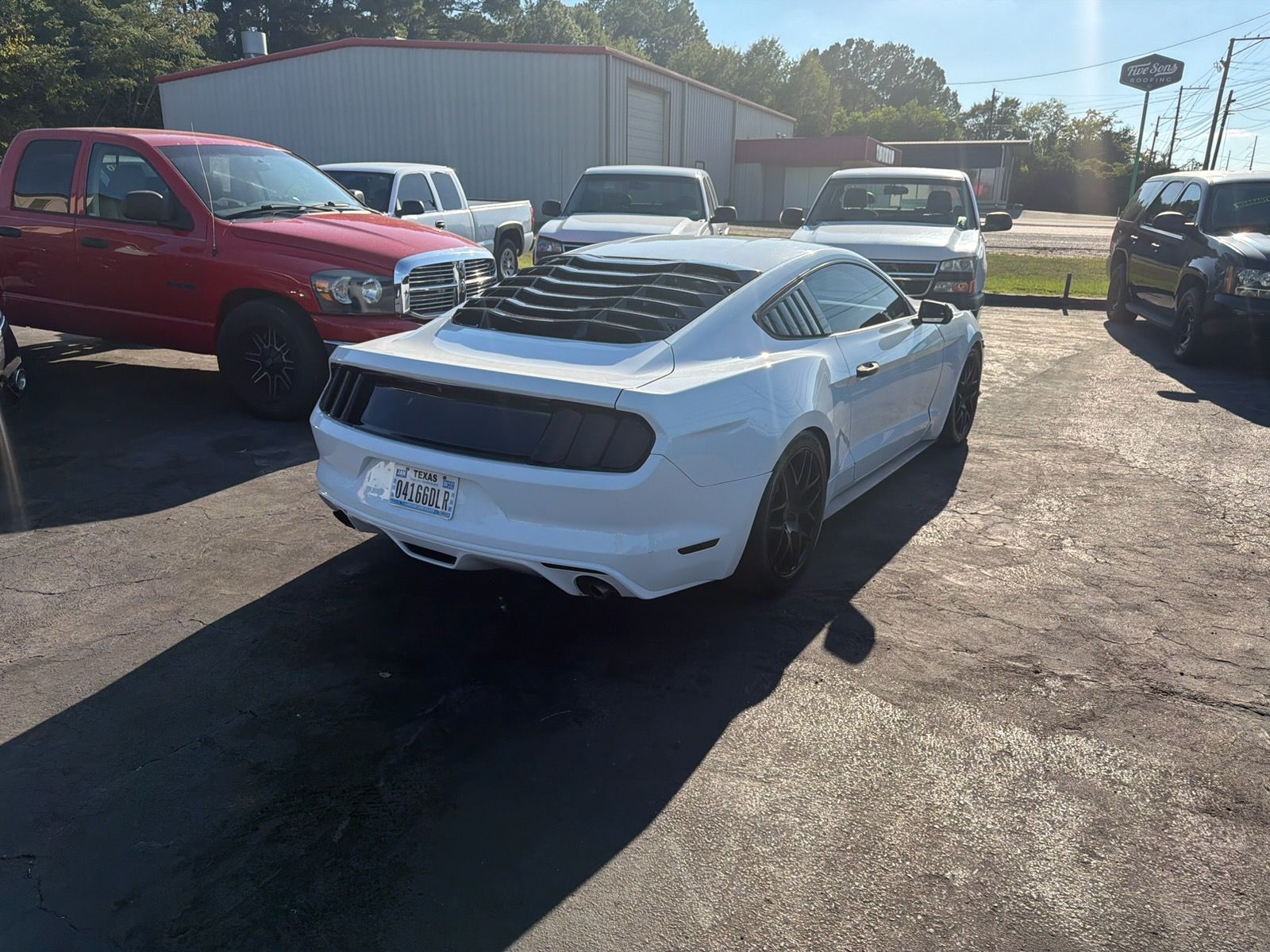 White sports car in a gravel lot with red and white trucks parked nearby under bright sunlight.