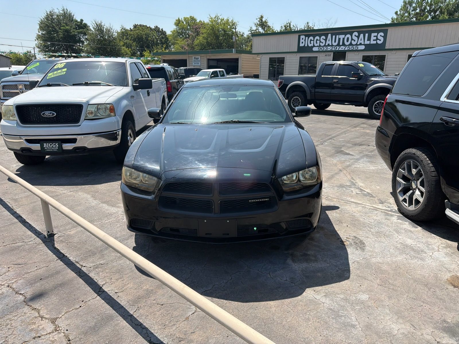 Black sedan parked in a used car lot beside white and black trucks, with a dealership building in the background