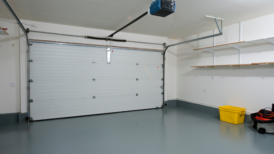 An empty garage with a white sectional door, gray epoxy floor, wall-mounted shelves, and a yellow plastic storage bin.
