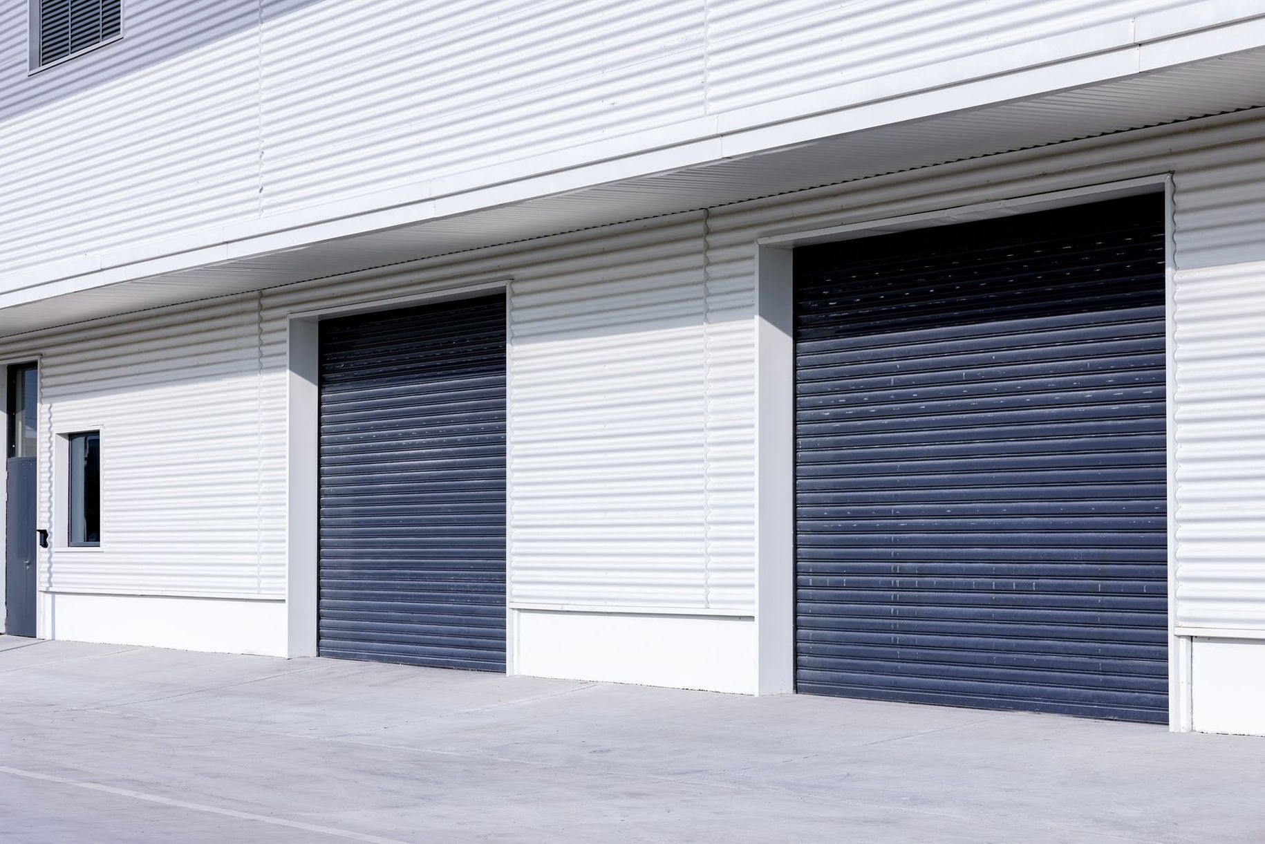 A white industrial building exterior with two large, closed dark gray roll-up metal bay doors.
