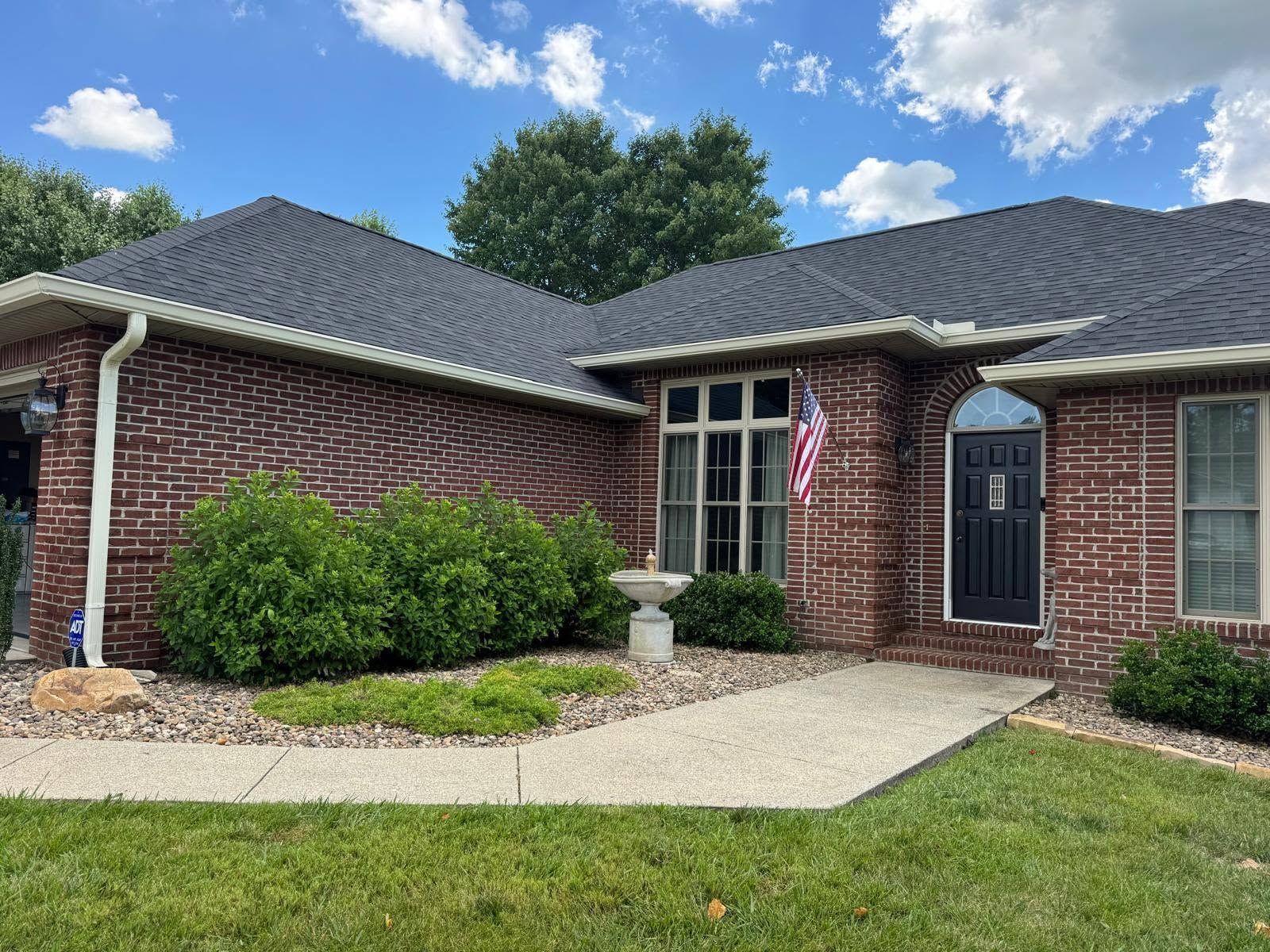Brick house with dark roof, front window, black door, and a gravel walkway by a green lawn.
