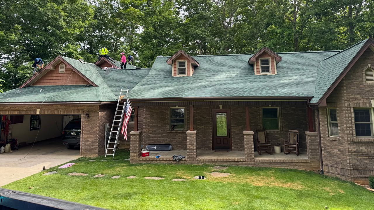 Brick house with gray roof and dormer windows, workers on the roof, and a ladder by the garage.
