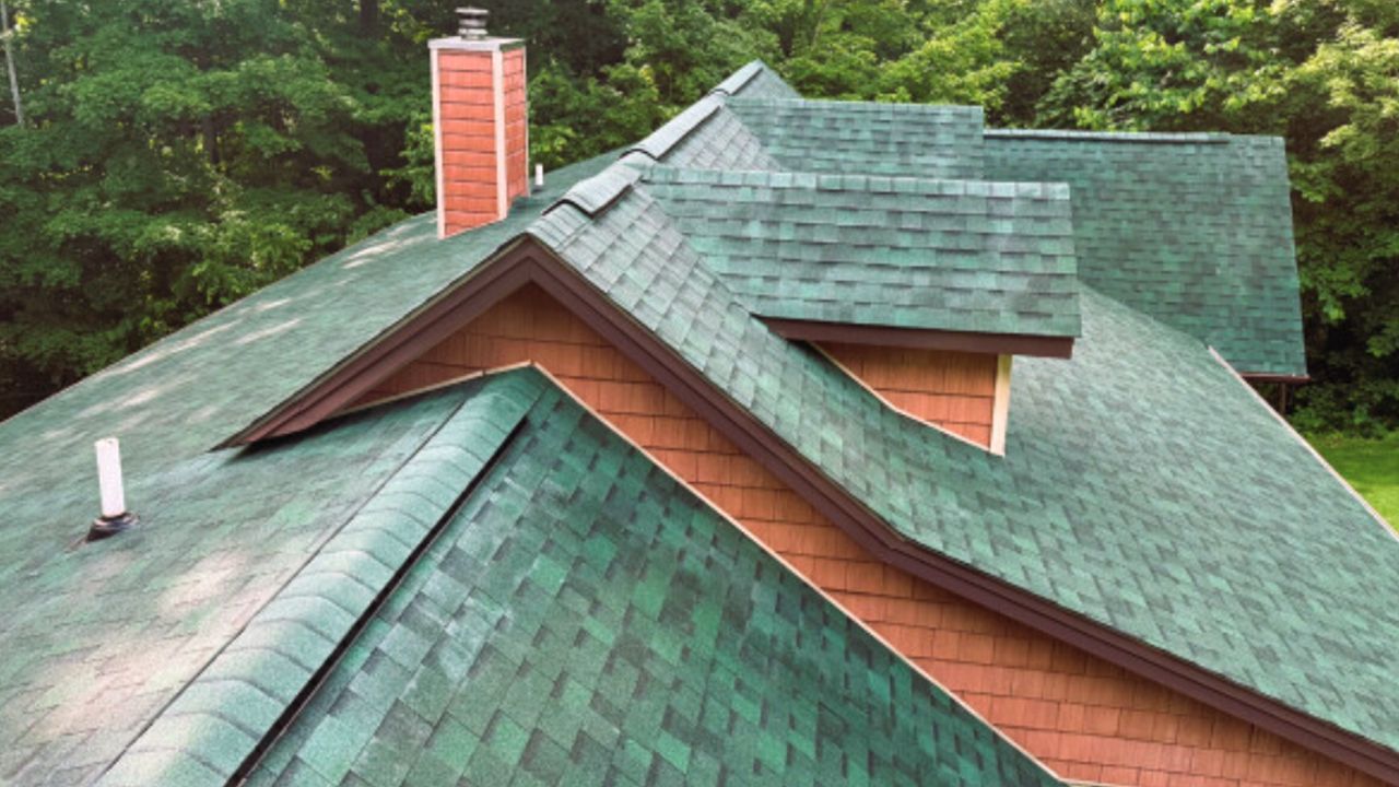 Green shingled house roof with dormers and red brick chimney amid trees