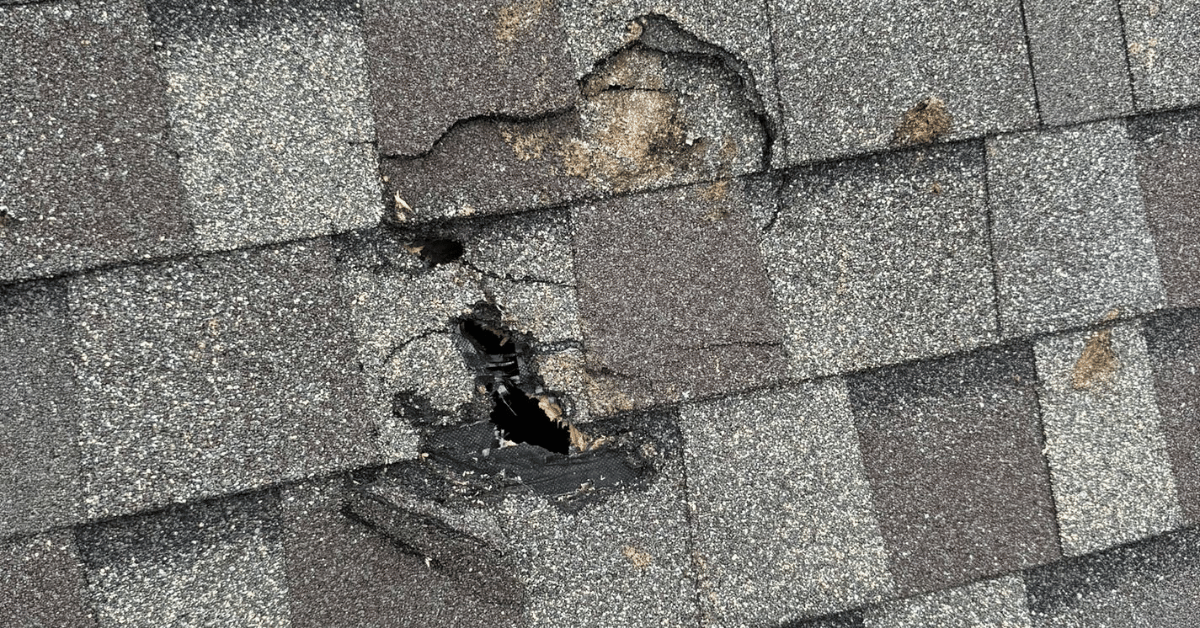 Damaged gray roof shingles with a large hole and missing pieces