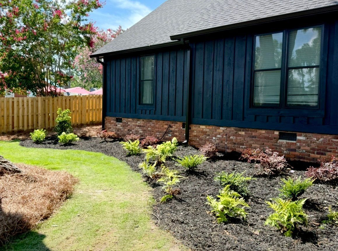 A house with navy blue siding and black-framed windows. A garden bed with mulch and green plants lines the house.