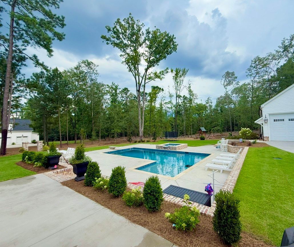 Backyard pool with a stone patio and landscaping, featuring a light blue pool surrounded by greenery and a white garage.