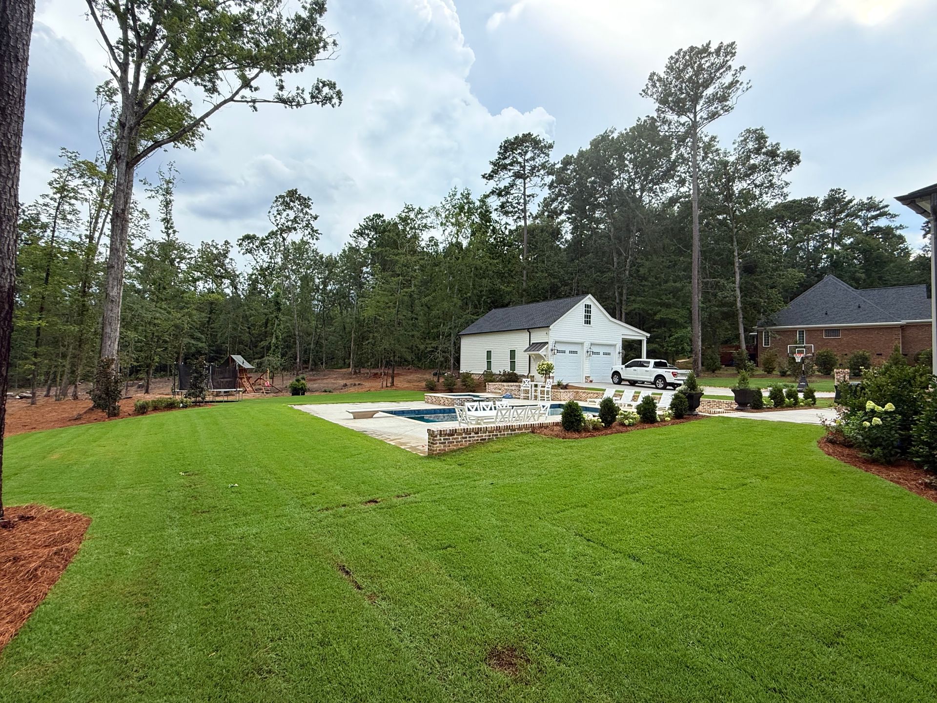 A backyard with a pool, garage, and lush green lawn. A car is parked in front of the garage, and trees surround the property.