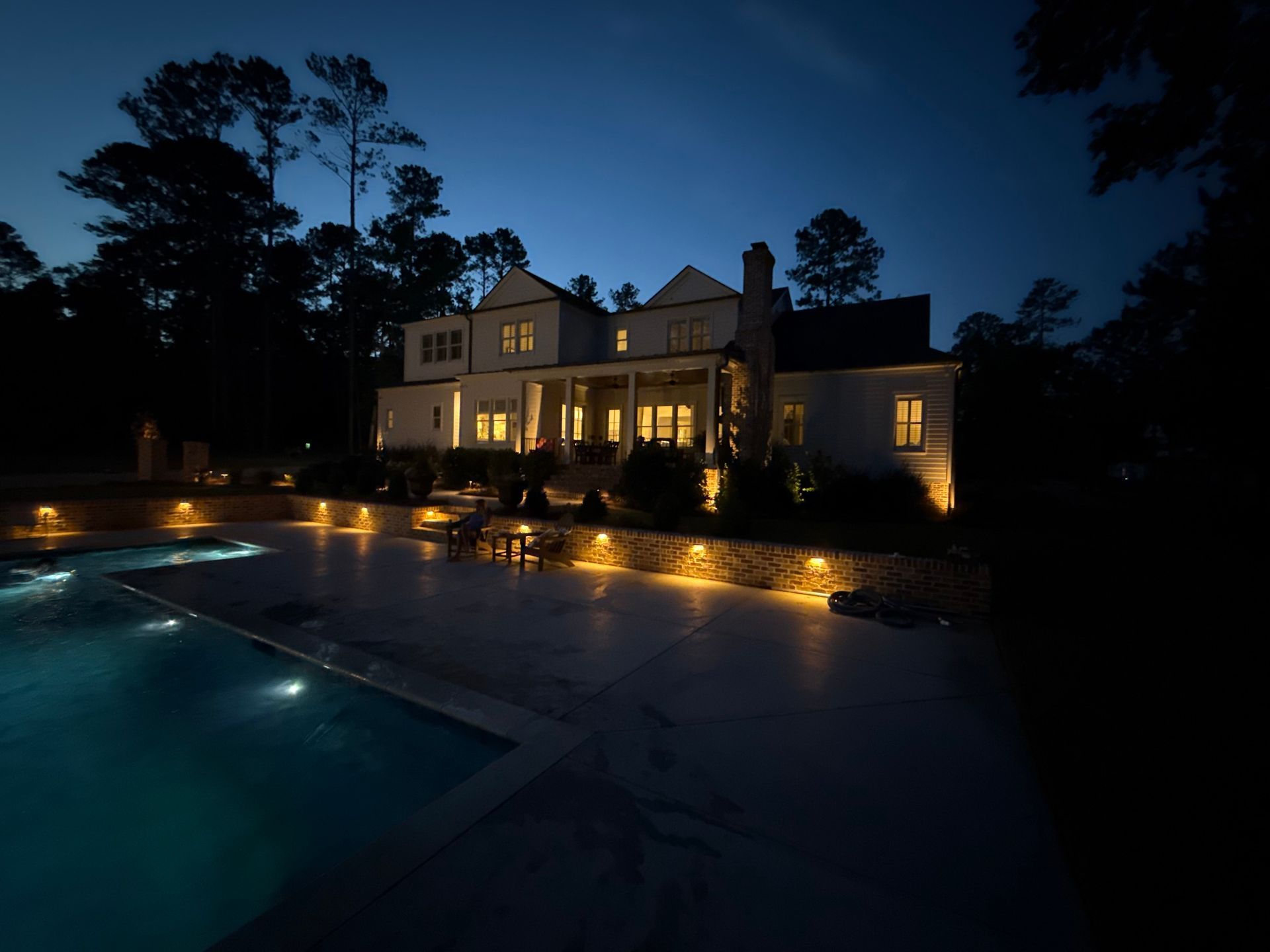 A large, lit-up house at night with a pool in the foreground, surrounded by landscape lighting.
