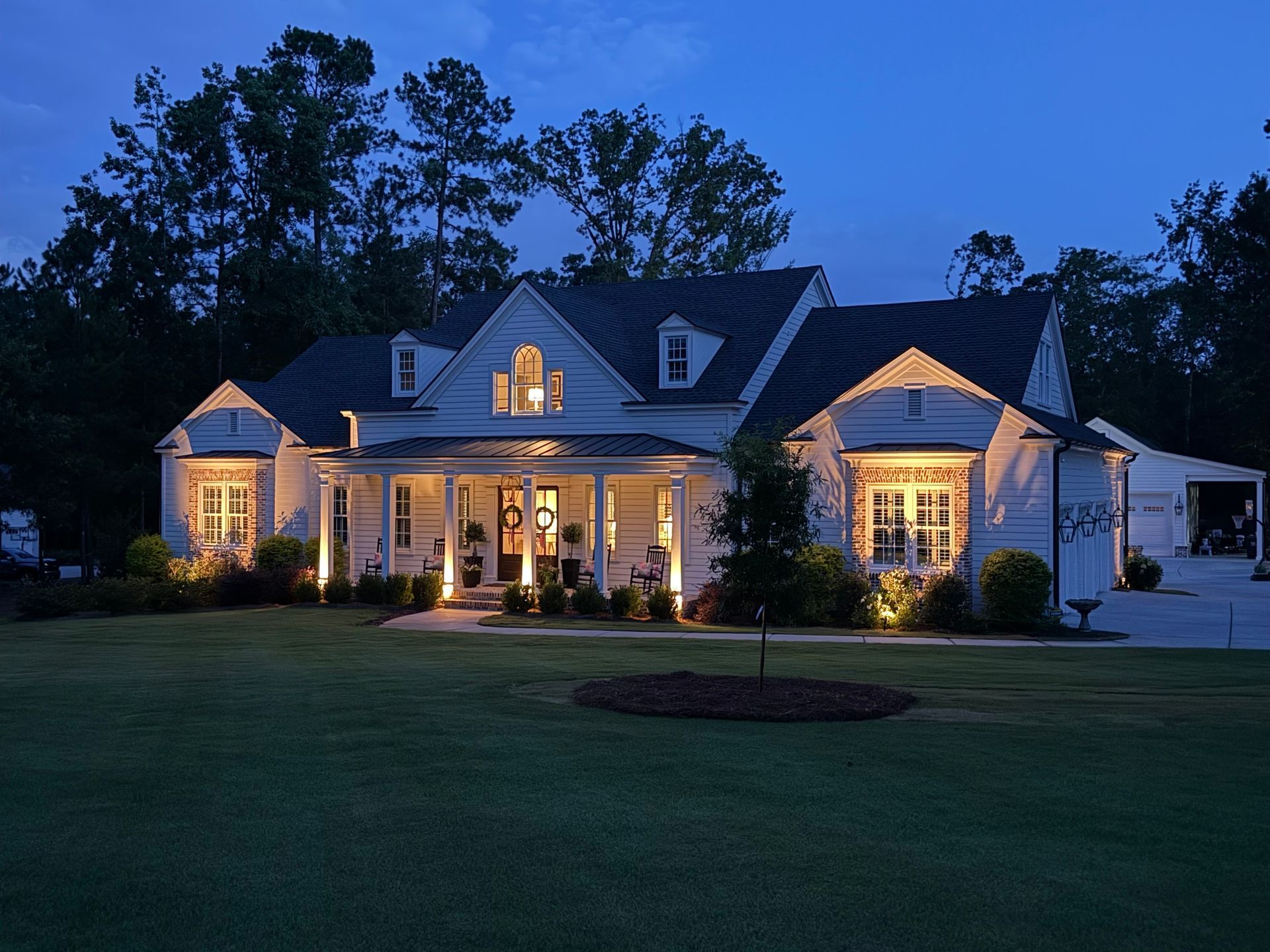 White house lit with outdoor lights at dusk, on a large lawn. Trees in the background.
