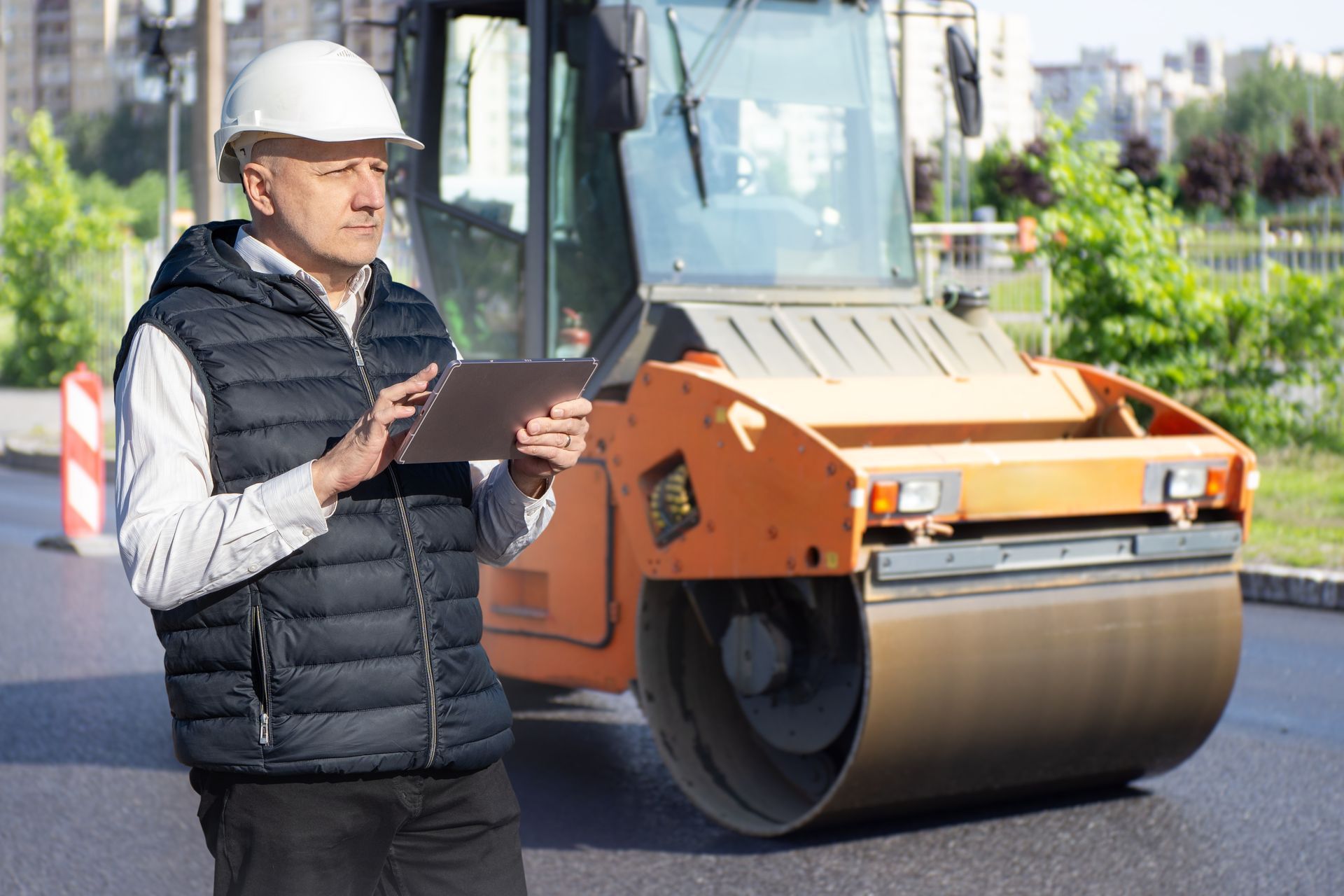Construction worker in hard hat and vest holding a tablet, road roller in background. Construction worker in hard hat and vest holding a tablet, road roller in background.