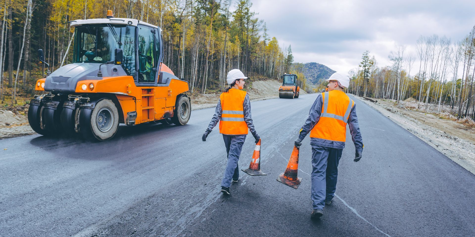 Two workers in orange vests walk on a newly paved road, with construction equipment in the background. Two workers in orange vests walk on a newly paved road, with construction equipment in the background.