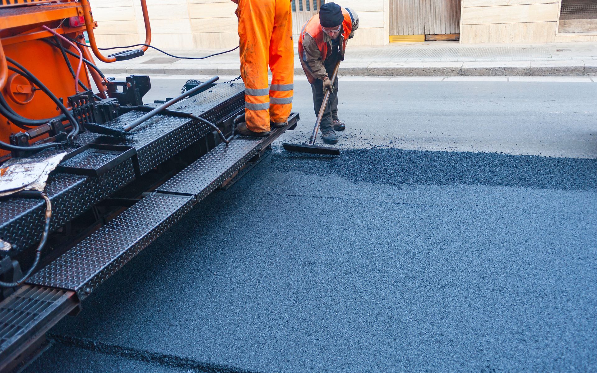 Workers paving a road with asphalt; one rakes, the other stands by the machine in orange overalls. Workers paving a road with asphalt; one rakes, the other stands by the machine in orange overalls.