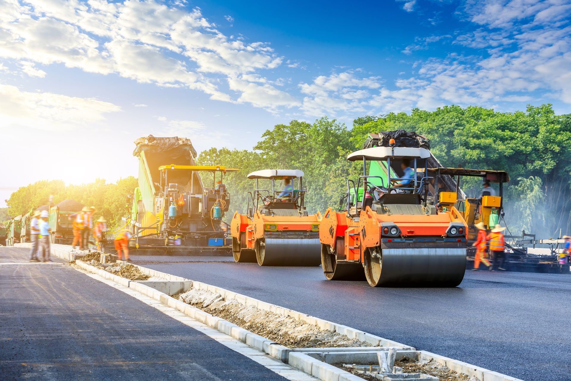 Road paving in progress, with asphalt machinery and workers in orange vests on a sunny day. Road paving in progress, with asphalt machinery and workers in orange vests on a sunny day.