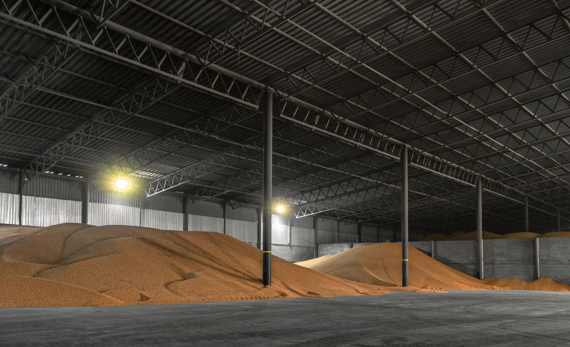 Large warehouse interior with piles of grain, dark metal beams, and interior lights.