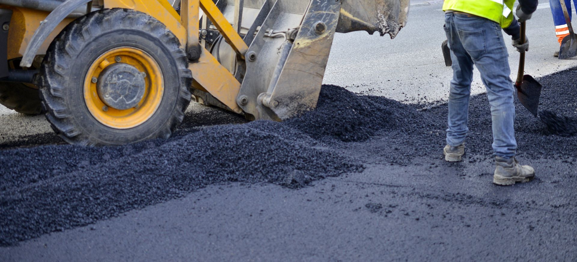 A yellow construction vehicle and a person shoveling asphalt onto a road during road work. A yellow construction vehicle and a person shoveling asphalt onto a road during road work.