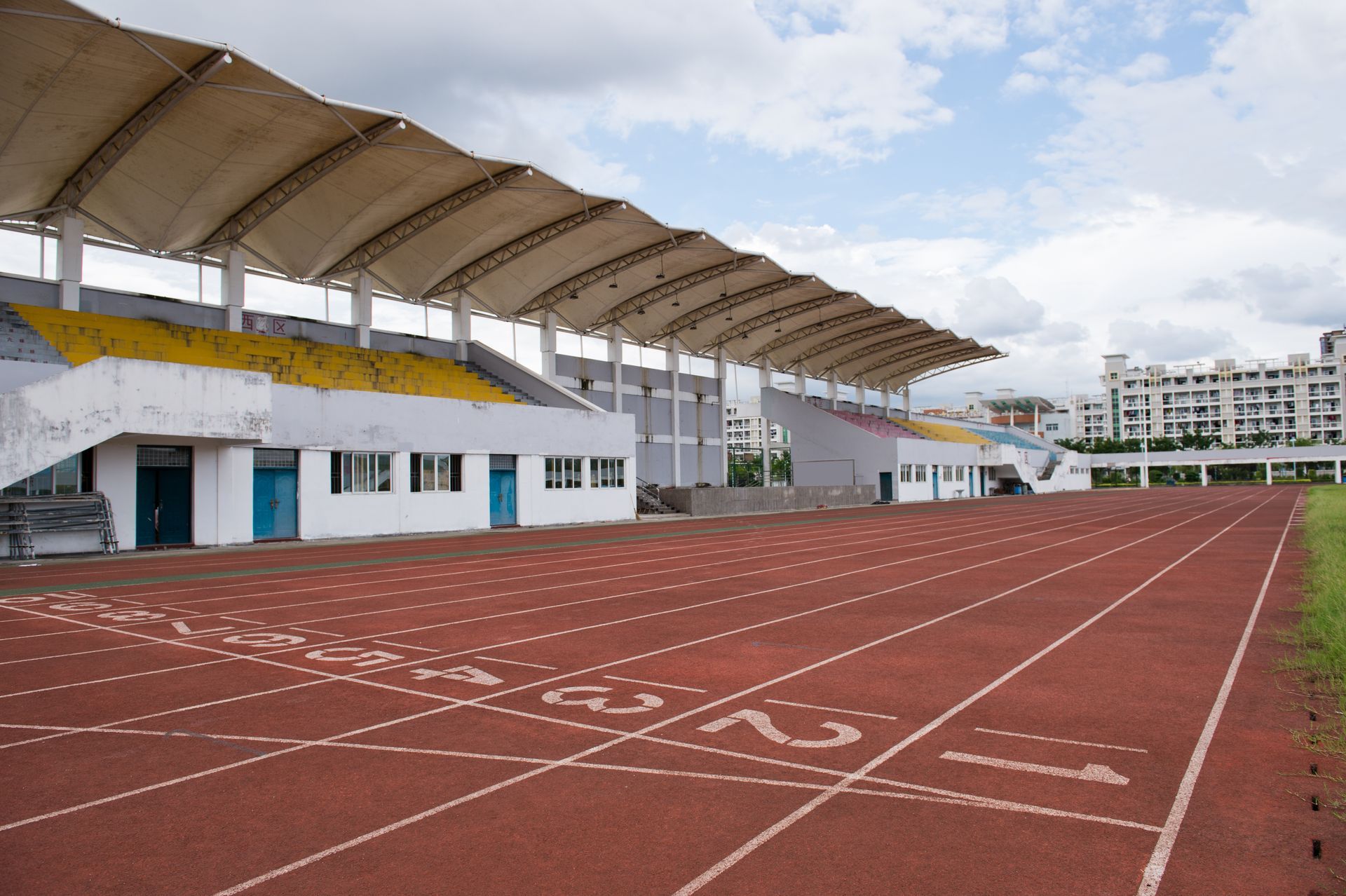 Red track at an outdoor stadium with white and yellow seating under a canopy.